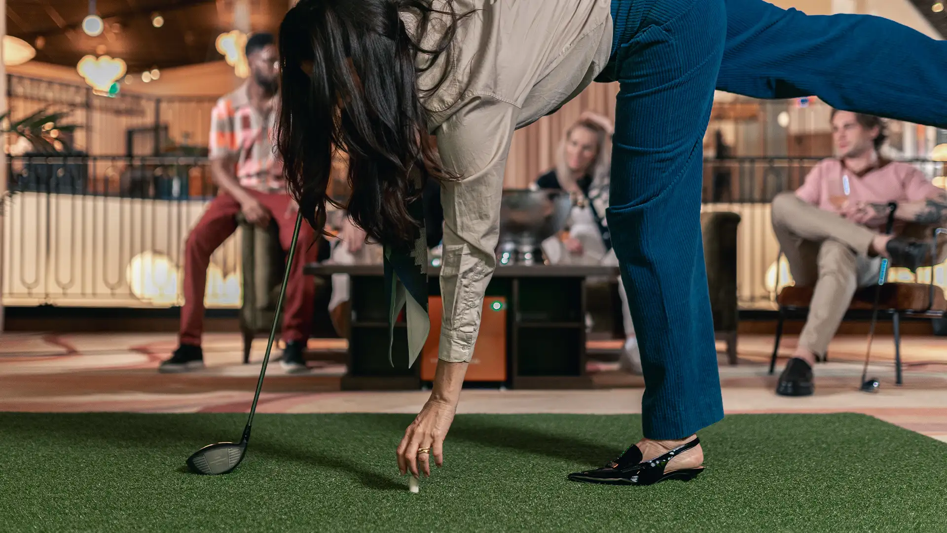 Kvinna förbereder ett slag i inomhusgolf medan vänner ser på inne i en elegant lounge-miljö.