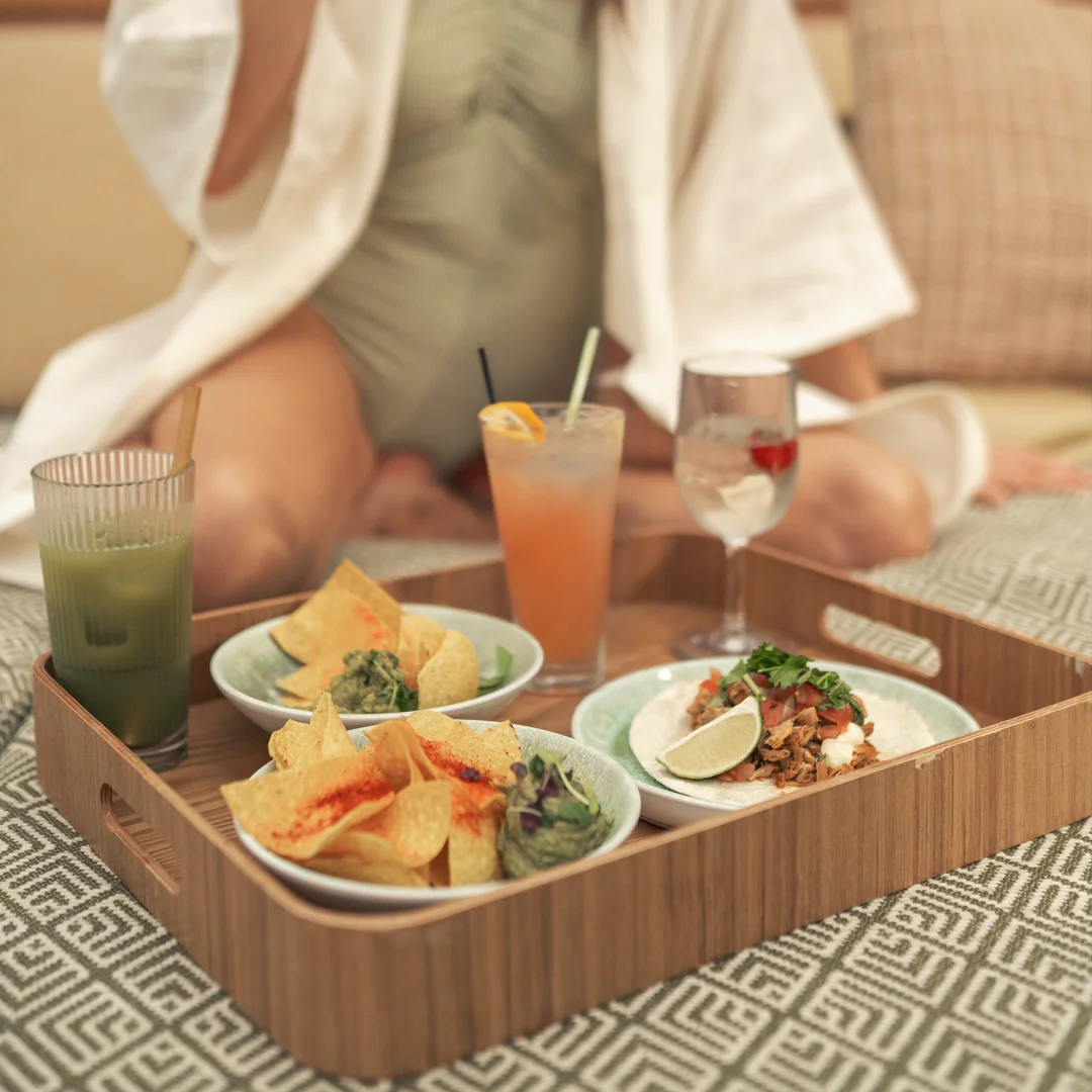 Bricka med nachos, smårätter och färgglada drinkar serveras vid poolen på Pachanga Spa i Göteborg. Kvinna i baddräkt och morgonrock i bakgrunden.