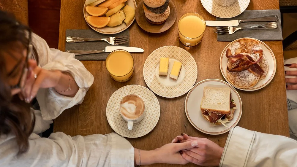 Två kvinnor håller varandra i handen över en hotellfrukost på Archie’s efter en morgon på spa. Pannkakor, smörgåsar, bacon och kaffe står på bordet.